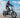 A rider cruising on a sleek black WILDEWAY FW11 folding fat-tire electric bike across a sunny, soft sandy beach next to the ocean waves.